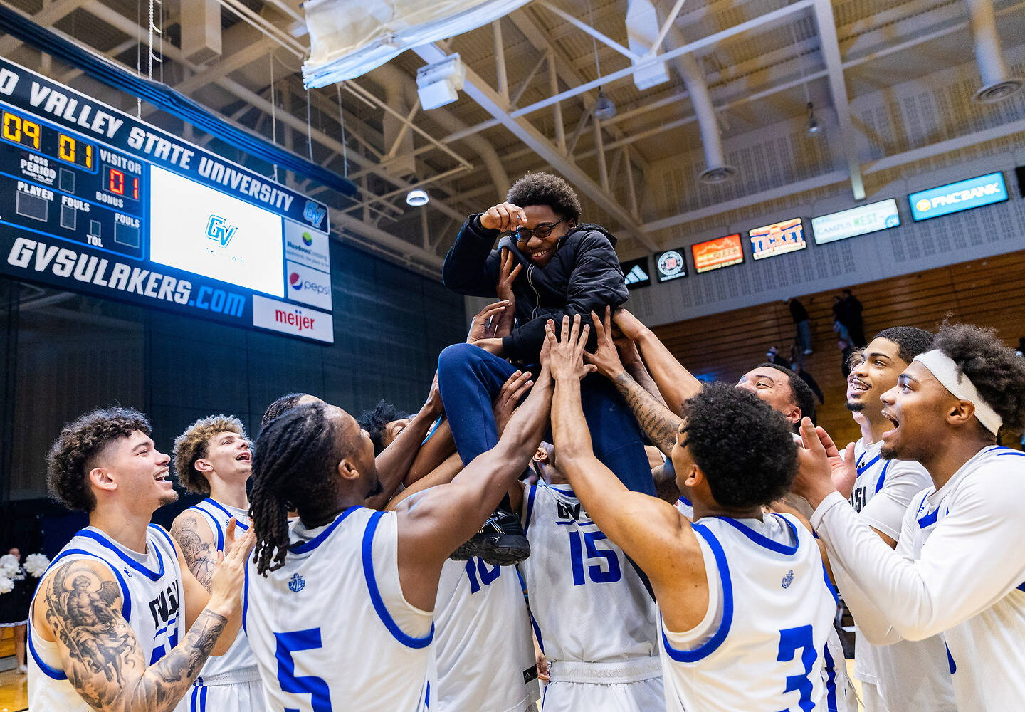 A group of basketball players in white and blue uniforms joyfully lifts a teammate into the air during a celebratory moment on the court.